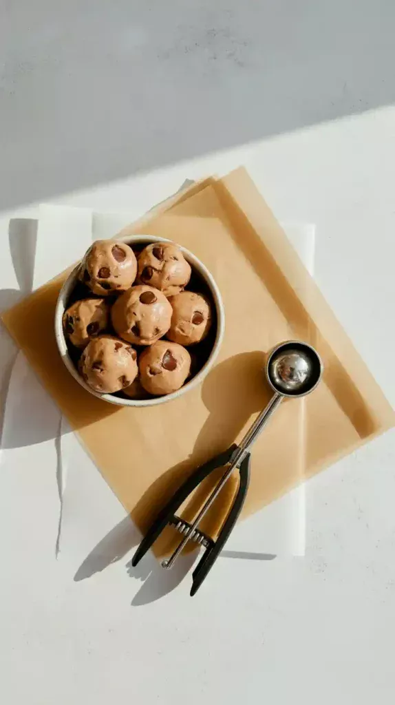 A flat lay image showing chilled balls of cookie dough, a cookie scoop, and a piece of parchment paper for making air fryer cookies.