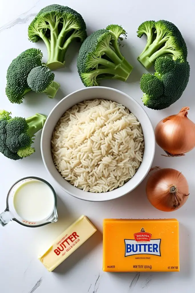 A bright and clean flat lay of ingredients for broccoli cheese rice casserole: fresh broccoli florets, uncooked rice, a block of cheddar cheese, milk, butter, and an onion on a white background