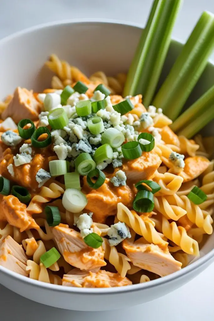  A bowl of buffalo chicken pasta garnished with blue cheese crumbles and green onions, with a side of celery sticks.