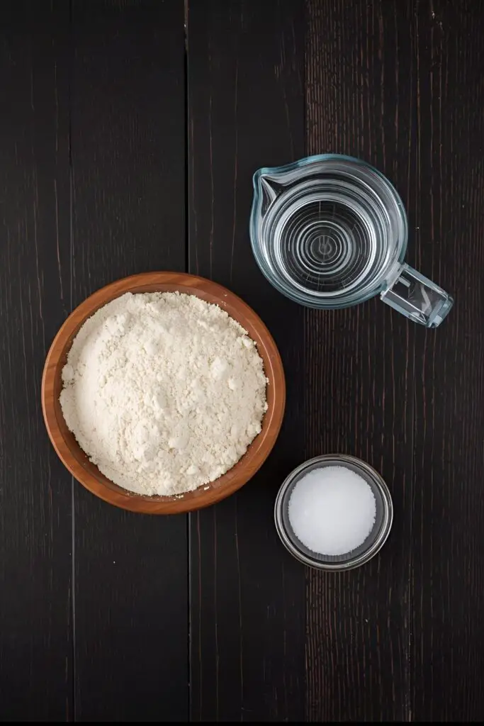 A flat-lay collage of chapati ingredients: a bowl of atta flour, a measuring cup of water, and a small dish of salt on a wooden surface.