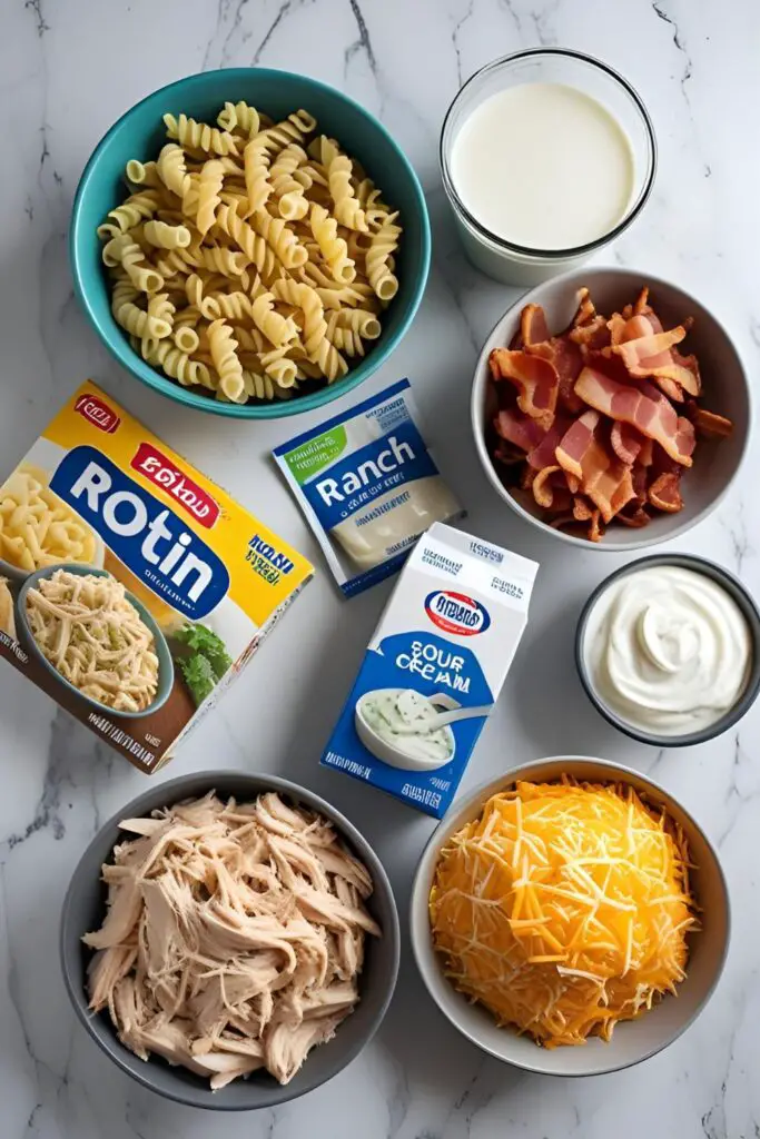 A flat lay of the ingredients for chicken bacon ranch casserole, including pasta, shredded chicken, crispy bacon, ranch seasoning, milk, and cheddar cheese, neatly arranged on a countertop.