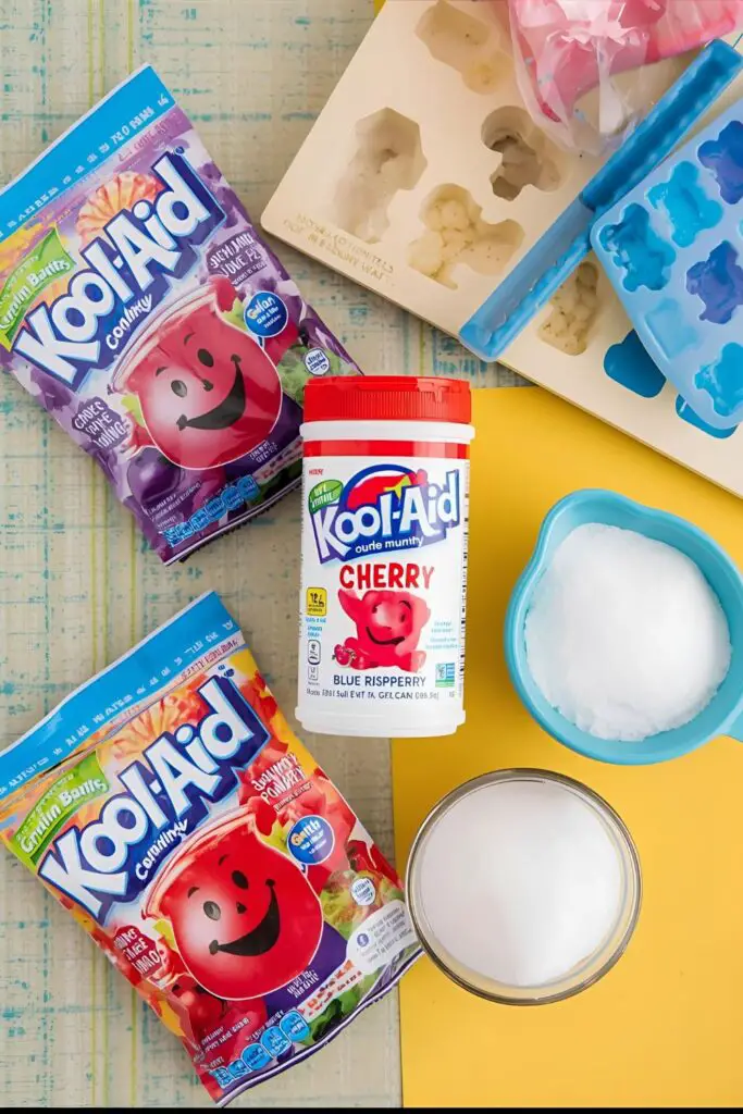 A flat lay of ingredients for Kool-Aid gummy bears: unflavored gelatin, Kool-Aid packets, water, and sugar.