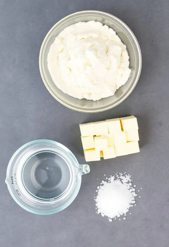 A neat flat lay collage of the essential pastry dough ingredients: a bowl of flour, a block of very cold butter cut into cubes, a measuring cup of ice water, and a sprinkle of salt.