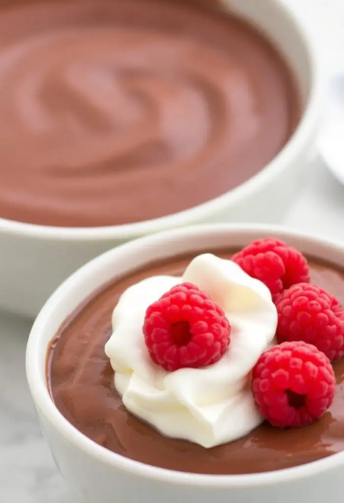 A close-up shot showing two different servings of chocolate pudding. One is a simple, classic bowl of smooth pudding. The other is a more decadent version topped with a generous dollop of whipped cream and fresh raspberries.