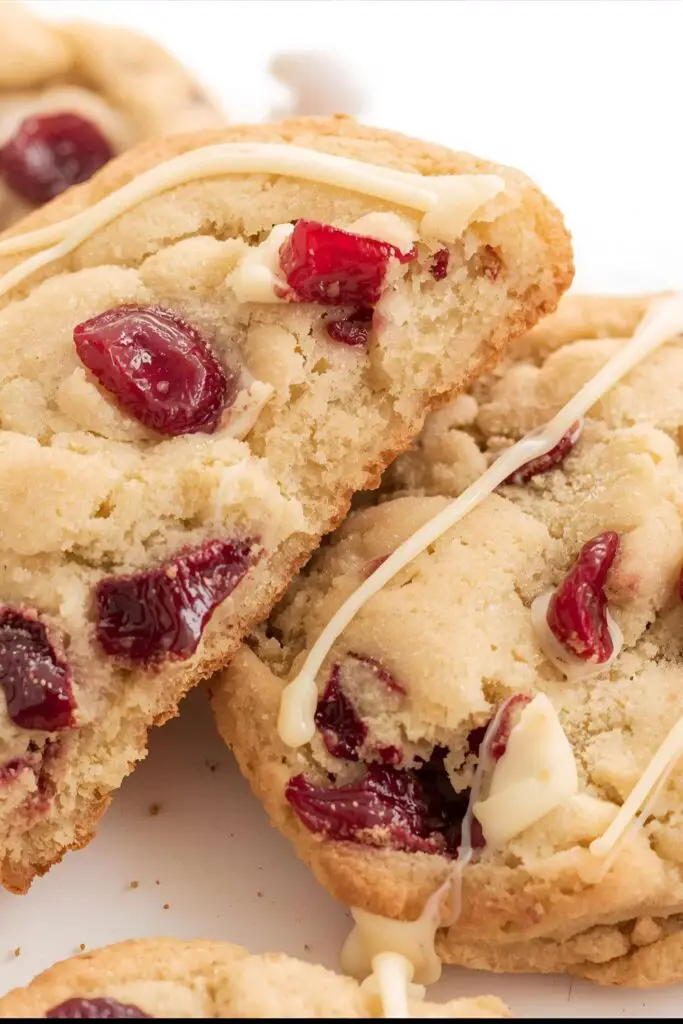 A close-up shot of two white chocolate cranberry cookies. One is whole, showing its perfectly golden edges, and the other is broken in half to reveal the soft, chewy interior with melted white chocolate and vibrant cranberries.
