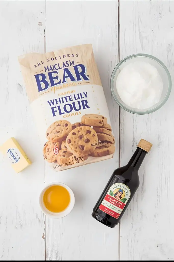 A clean, Southern-inspired flat lay of ingredients for White Lily cookies: the iconic flour bag, a stick of butter, powdered sugar, an egg yolk, and a bottle of vanilla on a whitewashed wood background.