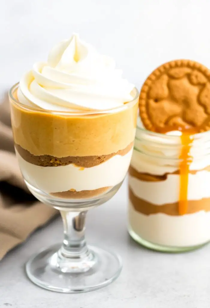 A close-up shot of two beautifully layered pumpkin cheesecake parfaits. One is in a simple, elegant glass, topped with a perfect swirl of whipped cream. The other is in a small mason jar, garnished with a whole gingersnap cookie and a drizzle of caramel.
